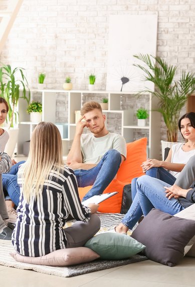 A group of seven people is sitting in a modern room on bean bags, engaging in discussion. Plants and shelves are visible in the background.