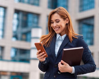 Eine lächelnde Frau in einem Anzug steht vor einem modernen Gebäude und schaut auf ihr Smartphone, während sie eine Mappe in der anderen Hand hält.