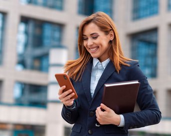 Eine lächelnde Frau in einem Anzug steht vor einem modernen Gebäude und schaut auf ihr Smartphone, während sie eine Mappe in der anderen Hand hält.