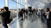Besucher stehen in einem modernen Raum mit großen Fenstern, die einen Blick auf die Stadt bieten. Einige Personen fotografieren die Aussicht, während andere die Szenerie betrachten. Die Wände sind mit blauen Fliesen dekoriert.
