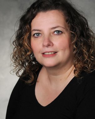 Portrait of a woman with curly brown hair, smiling in a black top. The background is neutral and blurred.