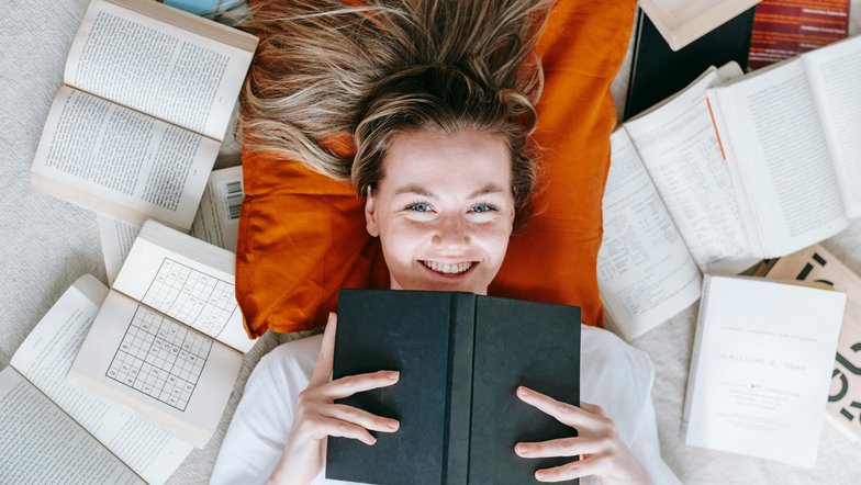Eine lächelnde Frau liegt auf einem orangefarbenen Kissen, umgeben von offenen und geschlossenen Büchern. Sie hält ein schwarzes Buch vor sich und schaut direkt in die Kamera.