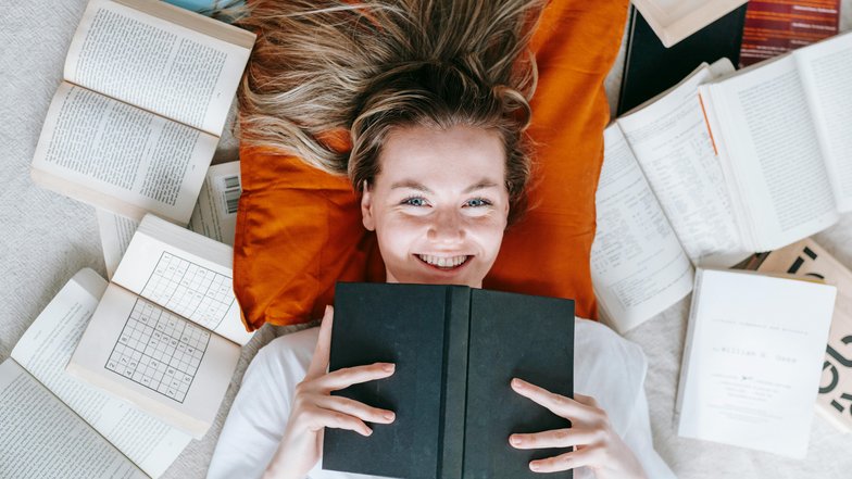 Eine lächelnde Frau liegt auf einem orangefarbenen Kissen, umgeben von offenen und geschlossenen Büchern. Sie hält ein schwarzes Buch vor sich und schaut direkt in die Kamera.