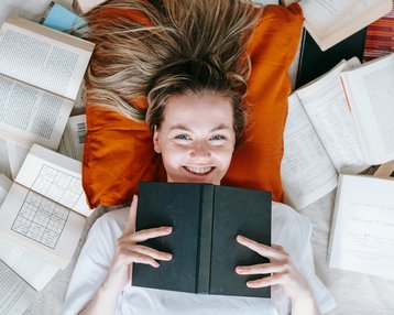 Kategorie Psychologie studieren Studium SRH University Eine lächelnde Frau liegt auf einem orangefarbenen Kissen, umgeben von offenen und geschlossenen Büchern. Sie hält ein schwarzes Buch vor sich und schaut direkt in die Kamera.