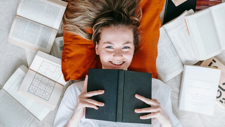 Eine lächelnde Frau liegt auf einem orangefarbenen Kissen, umgeben von offenen und geschlossenen Büchern. Sie hält ein schwarzes Buch vor sich und schaut direkt in die Kamera.