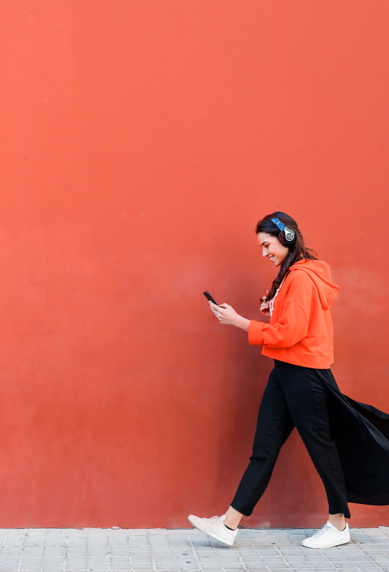 A woman in an orange hoodie walks along a red wall, looking at her smartphone.