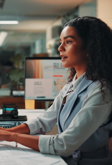Eine Frau mit lockigem Haar sitzt an einem Schreibtisch und arbeitet am Computer. Mehrere Monitore und Büromaterialien sind sichtbar.