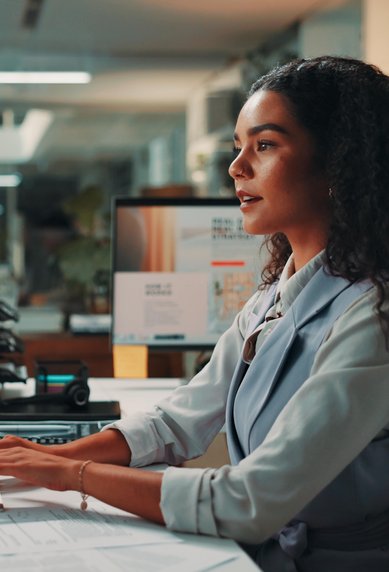 Eine Frau mit lockigem Haar sitzt an einem Schreibtisch und arbeitet am Computer. Mehrere Monitore und Büromaterialien sind sichtbar.