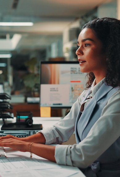 A woman with curly hair is sitting at a desk, working on a computer. Several monitors and office supplies are visible.