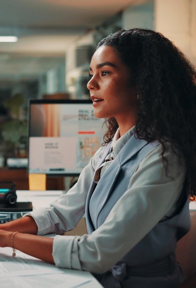 A woman with curly hair is sitting at a desk, working on a computer. Several monitors and office supplies are visible.