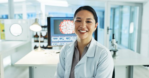 Eine Wissenschaftlerin in einem Labor lächelt, trägt einen weißen Laborkittel und sitzt vor einem Computer mit wissenschaftlichen Grafiken.