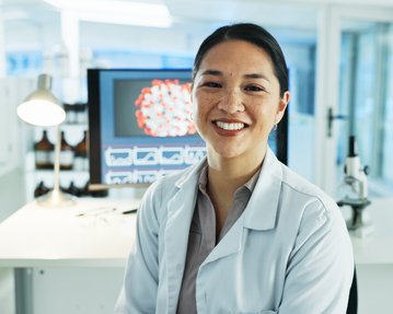 A scientist in a lab smiles, wearing a white lab coat, and sits in front of a computer displaying scientific graphs.