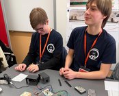 Zwei Jugendliche in T-Shirts arbeiten an einem Tisch mit elektronischen Komponenten, Kabeln und Baukästen.