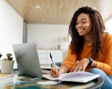 Eine lächelnde Frau mit lockigem Haar sitzt an einem Tisch, schreibt in ein Notizbuch und arbeitet an einem Laptop.