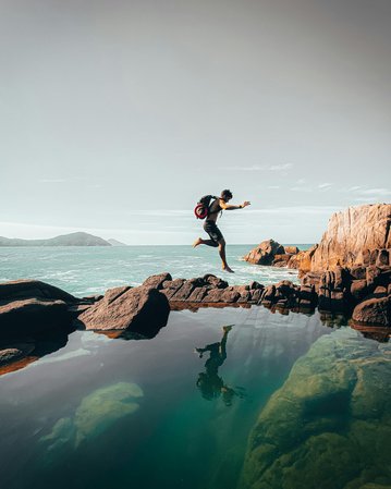 Eine Person springt von einem Felsen ins Wasser, umgeben von einer Küstenlandschaft mit Bergen im Hintergrund.