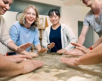 Eine Gruppe von sechs Personen steht um einen Tisch und klatscht mit den Händen auf die Tischoberfläche. Alle wirken fröhlich und engagiert in einer gemeinsamen Aktivität.