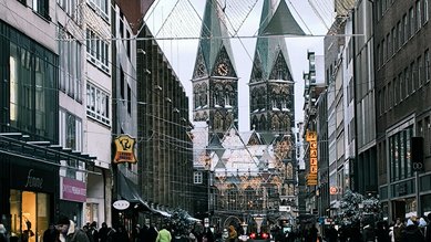 Blick auf eine belebte Straße mit Geschäften, im Hintergrund die markanten Türme einer Kirche und festliche Dekorationen.