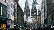 Blick auf eine belebte Straße mit Geschäften, im Hintergrund die markanten Türme einer Kirche und festliche Dekorationen.