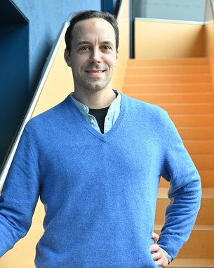 Ein Mann in einem blauen Pullover steht an einer Treppe mit orangefarbenen Stufen und lächelt freundlich.