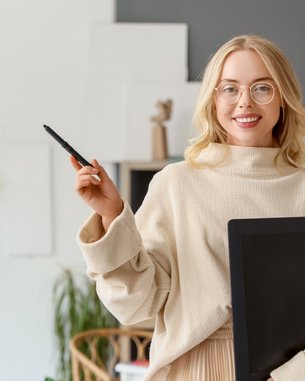 Eine lächelnde Frau mit Brille trägt einen beigen Pullover und hält einen Ordner in der Hand. Sie steht in einem kreativen Arbeitsumfeld mit einem Schreibtisch und bunten Notizen im Hintergrund.