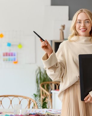 Eine lächelnde Frau mit Brille trägt einen beigen Pullover und hält einen Ordner in der Hand. Sie steht in einem kreativen Arbeitsumfeld mit einem Schreibtisch und bunten Notizen im Hintergrund.