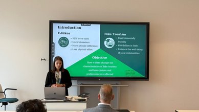 Eine Frau präsentiert Informationen über E-Bikes und Fahrradtourismus auf einem Bildschirm. Der Bildschirm zeigt Statistiken und Ziele der Präsentation, während Zuhörer im Hintergrund aufmerksam folgen.
