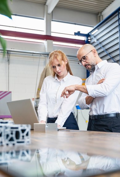 Two people in a modern office are discussing information on a laptop while standing at a table with technical components.