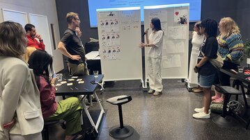 A group of people stands around two presentation boards while one person is giving a presentation. Some participants are taking notes or photos.
