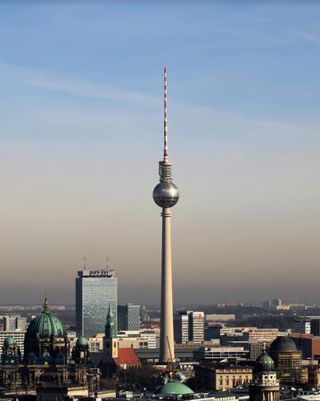 Fernsehturm Berlin ragt über die Stadt, umgeben von modernen Gebäuden und historischen Kirchen. Klarer Himmel im Hintergrund.
