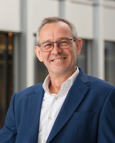 Ein Mann mit Brille und grauen Haaren trägt einen blauen Anzug und ein weißes Hemd. Er lächelt vor einem modernen Hintergrund.