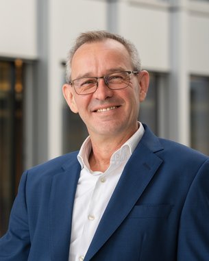 A man with glasses and gray hair is wearing a blue suit and a white shirt. He is smiling against a modern background.