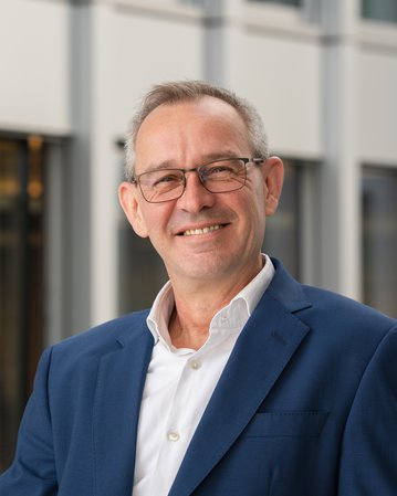 Ein Mann mit Brille und grauen Haaren trägt einen blauen Anzug und ein weißes Hemd. Er lächelt vor einem modernen Hintergrund.