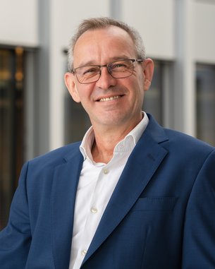 A man with glasses and gray hair is wearing a blue suit and a white shirt. He is smiling against a modern background.