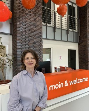 Eine Frau steht an einem Empfangstresen mit der Aufschrift "moin & welcome". Im Hintergrund hängen orangefarbene Lampen, und es gibt Pflanzen in Töpfen.