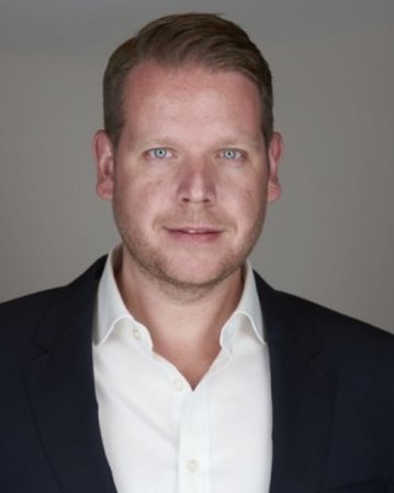 Mann mit kurzem, blondem Haar, trägt ein weißes Hemd und einen dunklen Blazer, schaut direkt in die Kamera.