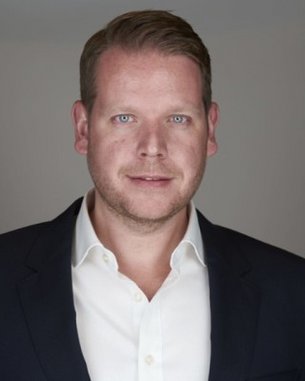 Mann mit kurzem, blondem Haar, trägt ein weißes Hemd und einen dunklen Blazer, schaut direkt in die Kamera.