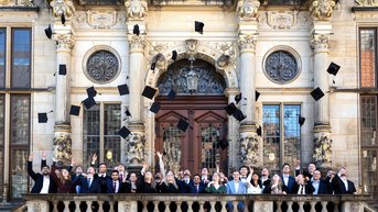 Gruppe von Absolventen, die auf einem Balkon stehen und ihre Hüte in die Luft werfen, vor einem historischen Gebäude.