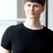 A person with short brown hair and glasses is wearing a black top. They are standing in front of a light background.
