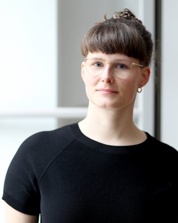 Eine Person mit kurzen, braunen Haaren und Brille trägt ein schwarzes Oberteil und schaut in die Kamera.