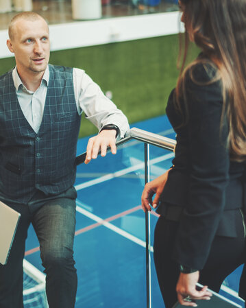 Two people in formal attire are having a conversation in a sports hall. One person is standing on a staircase while the other is standing below.