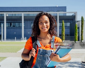 Eine junge Frau mit lockigem Haar trägt einen Rucksack und hält mehrere Hefte in der Hand, vor einem modernen Gebäude im Freien.