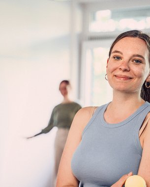 Eine lächelnde Frau in einem blauen Oberteil steht in einem hellen Raum. Im Hintergrund sind zwei Personen zu sehen, die miteinander interagieren.