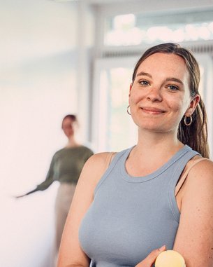 Eine lächelnde Frau in einem blauen Oberteil steht in einem hellen Raum. Im Hintergrund sind zwei Personen zu sehen, die miteinander interagieren.