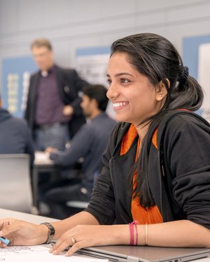Eine Studentin sitzt an einem Tisch und hält einen Stift in der Hand. Sie lächelt und schaut auf ein Blatt Papier mit Skizzen. Im Hintergrund sind weitere Personen in einem Klassenzimmer zu sehen.