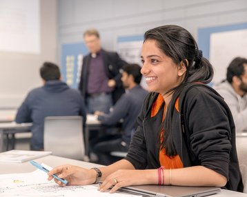 A student is sitting at a table, holding a pen in her hand. She is smiling and looking at a sheet of paper with sketches. In the background, other people can be seen in a classroom.
