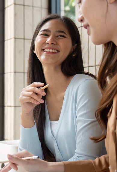 Zwei Frauen sitzen an einem Tisch, lächeln und unterhalten sich. Auf dem Tisch steht ein Laptop und Schreibmaterial.