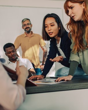 Eine Gruppe von fünf Personen diskutiert in einem modernen Raum. Zwei Frauen zeigen auf Dokumente auf einem Tisch, während ein Mann im Hintergrund zuschaut. Die Atmosphäre wirkt kollaborativ und engagiert.