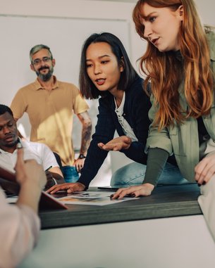 Eine Gruppe von fünf Personen diskutiert in einem modernen Raum. Zwei Frauen zeigen auf Dokumente auf einem Tisch, während ein Mann im Hintergrund zuschaut. Die Atmosphäre wirkt kollaborativ und engagiert.