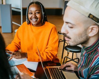 A person with braided hair is wearing an orange sweater and smiling while sitting at a table. In front of her are some documents, and another participant is working on a laptop.