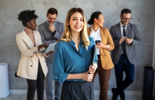 Eine lächelnde Frau in blauer Bluse hält einen Ordner, während vier Personen im Hintergrund miteinander kommunizieren und Geräte nutzen.
